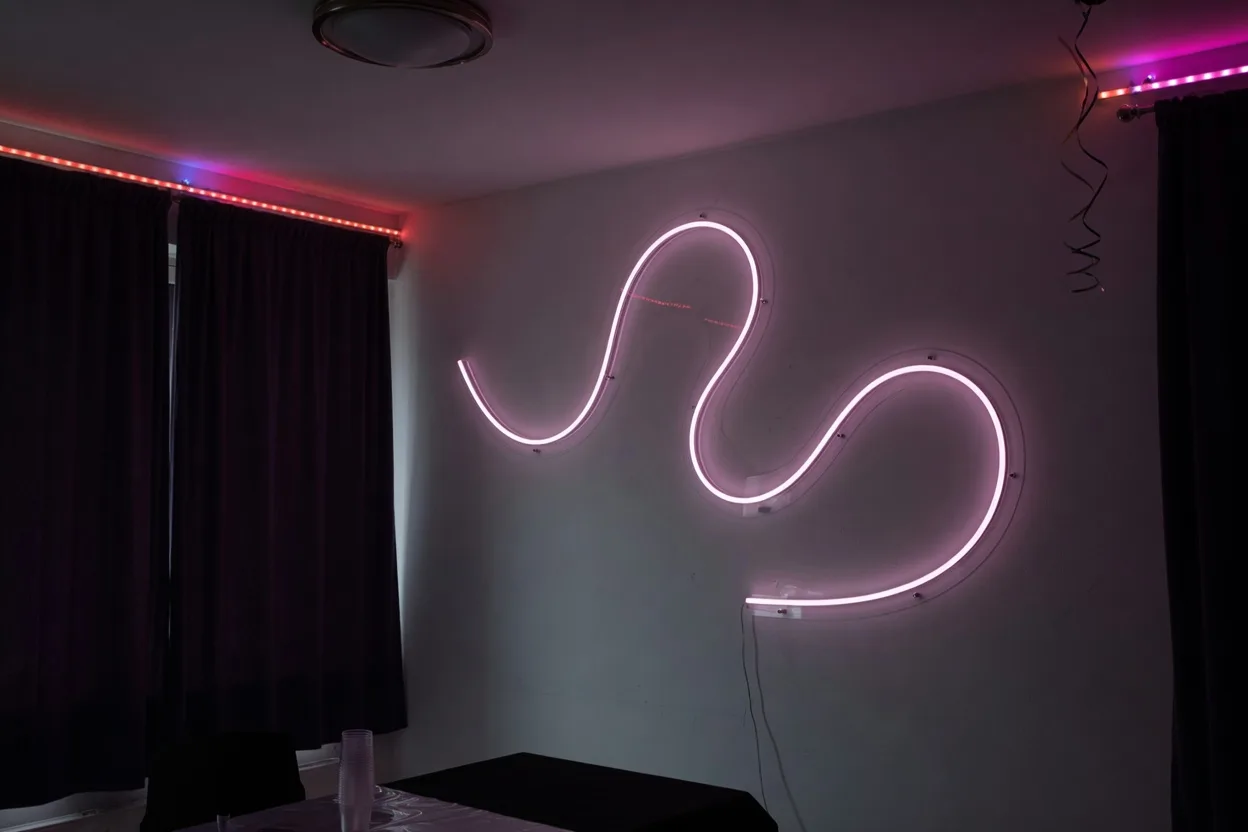 Salon préparé pour une fête, fenêtres occultées et plafonnier éteint, un néon LED sur un mur dégagé et des rubans LED colorés