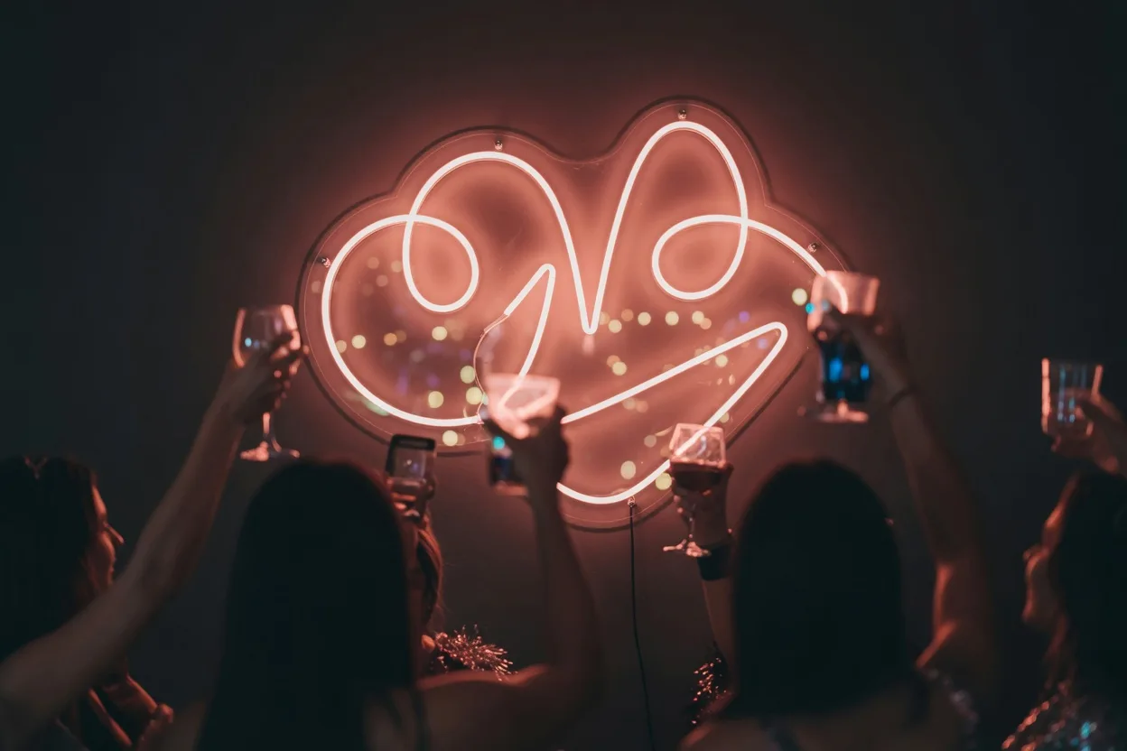 Coin photo d'une soirée devant un néon LED allumé sur un mur sombre, silhouettes floues d'invités levant des verres